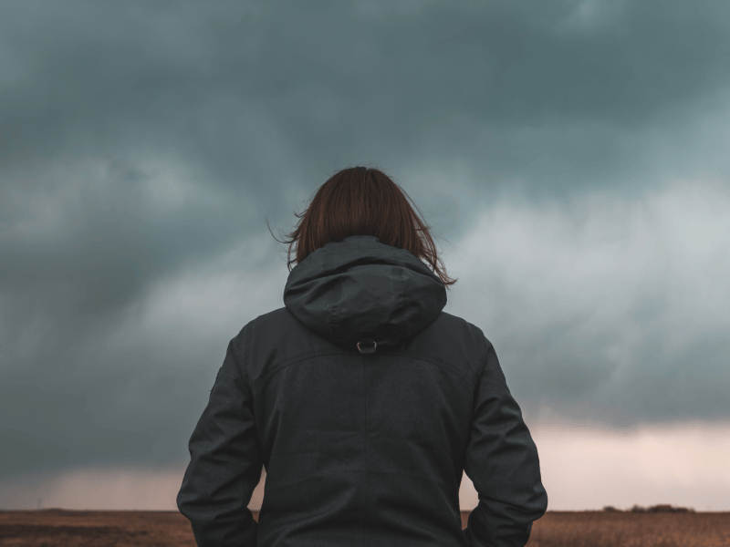 Frau steht mit dem Rücken zur Kamera vor einem düsteren Wolkenhimmel