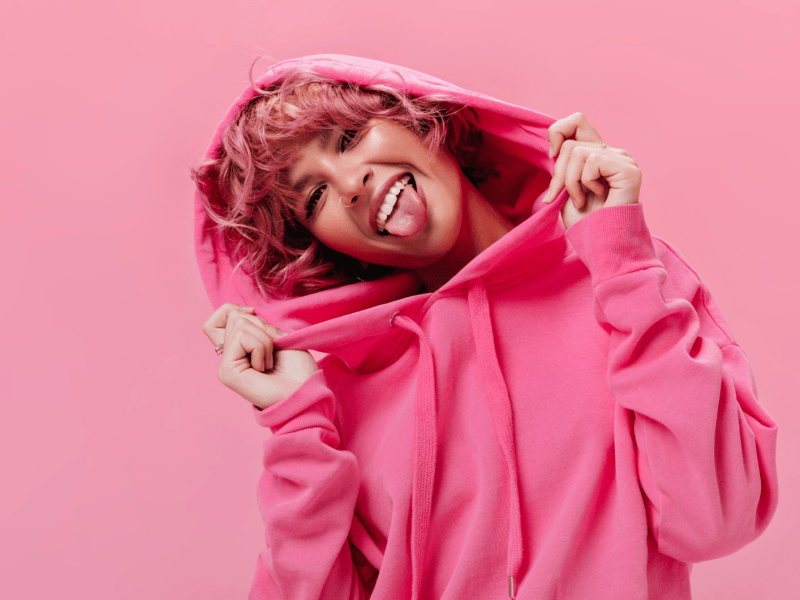 Frau mit pinken Kapuzenpulli, vor einer punken Wand mit pinken Haaren, die ihre Zunge herausstreckt und lächelt.