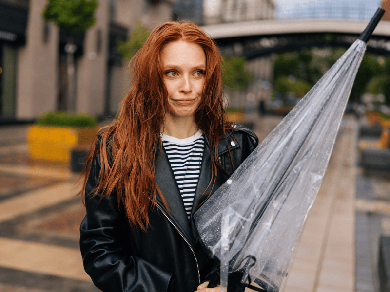 Frau mit nassen und durchwuschelten Haaren, die einen Regenschirm in der Hand hält und schmollend zur Seite schaut.