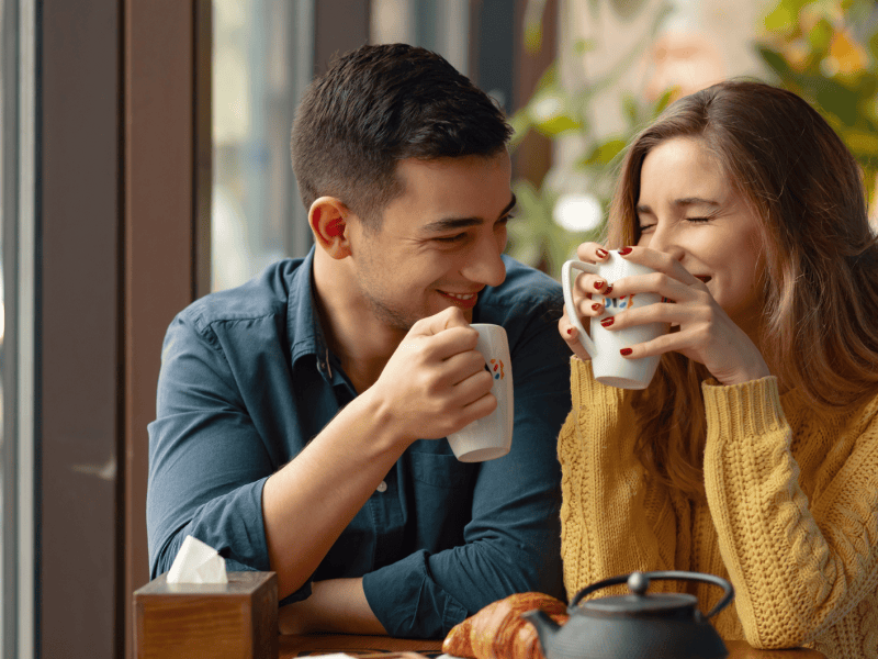 Frau und Mann mit Tassen in der Hand sitzen lachend an einem Tisch in einem Caf&eacute;