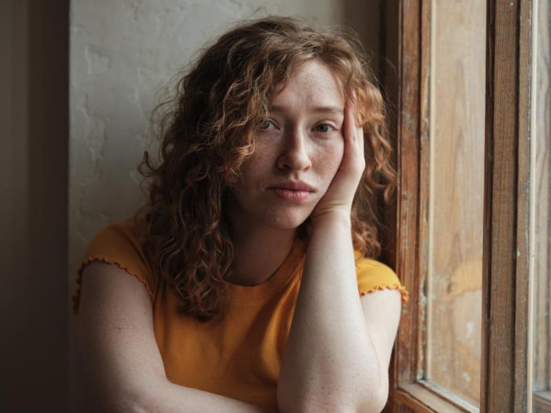 Junge Frau mit Locken sitzt am Fenster und guckt sehr traurig.