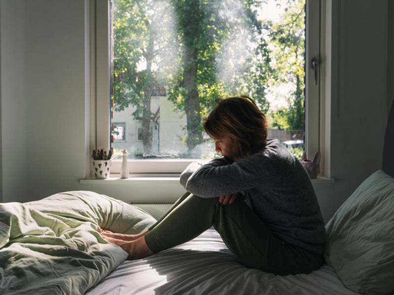Frau sitzt niedergeschlagen auf einem Bett vor einem Fenster.