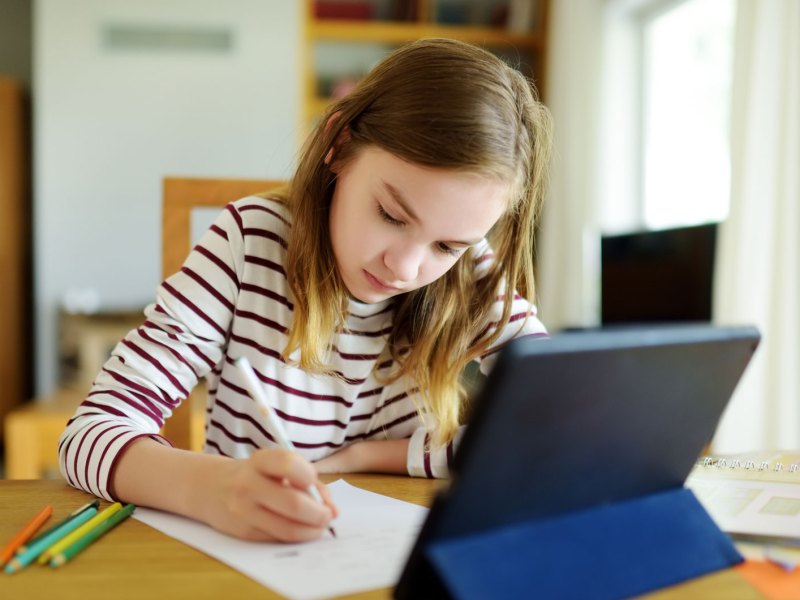 M&auml;dchen, ca. 9 Jahre alt, sitzt mit einem Tablet, Papier und Stiften am Tisch und erledigt Aufgaben.