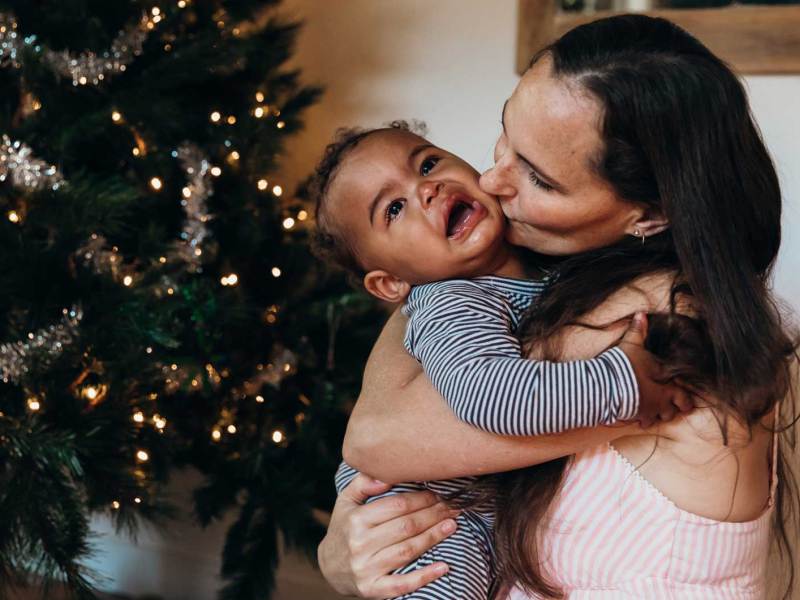 Mutter gibt ihrem weinenden Baby einen Kuss auf die Wange, während beide vor dem Weihnachtsbaum sitzen.