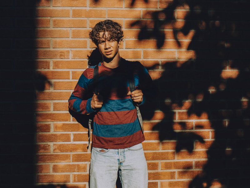 Junge im Teenageralter steht vor einer gemauerten Wand im Halbschatten und schaut ernst in Richtung Kamera. Er tr&auml;gt Jeans, Sweatshirt und Rucksack.
