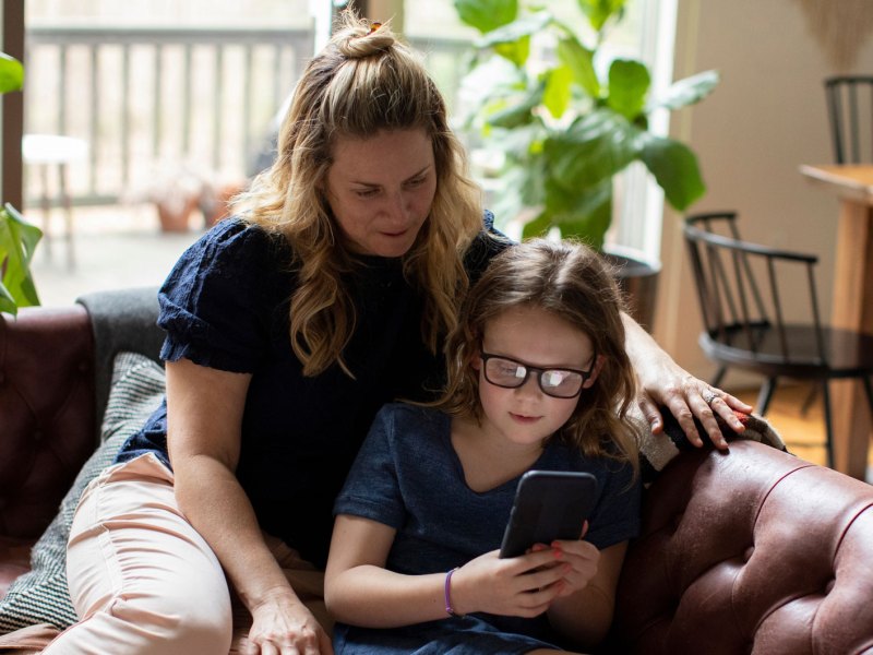 Mutter sitzt mit ihrer Tochter, ca. 8 Jahre alt, auf der Couch und schaut gemeinsam mit ihr aufs Smartphone.