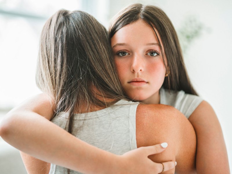 Besorgtes Teenager-M&auml;dchen umarmt ihre Mutter. Das M&auml;dchen schaut dabei direkt in die Kamera, die Mutter ist nur von hinten zu sehen.