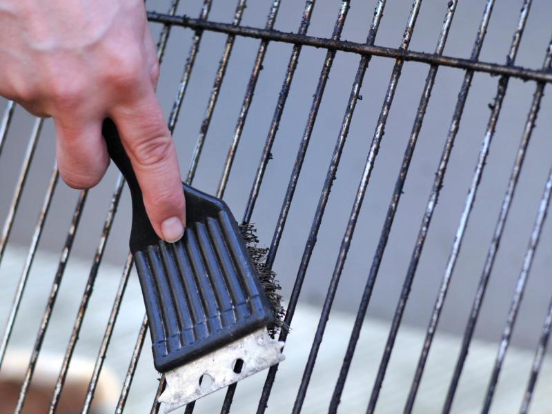 Eine Hand mit Grillb&uuml;rste reinigt einen stark verschmutzten Grillrost &uuml;ber hellem Boden im Freien bei Tageslicht.