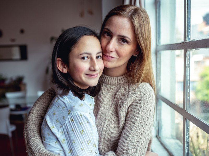 Mutter und Teenagertochter umarmen sich vor einem Fenster. Die Tochter lacht entspannt, die Mutter sieht besorgt aus.