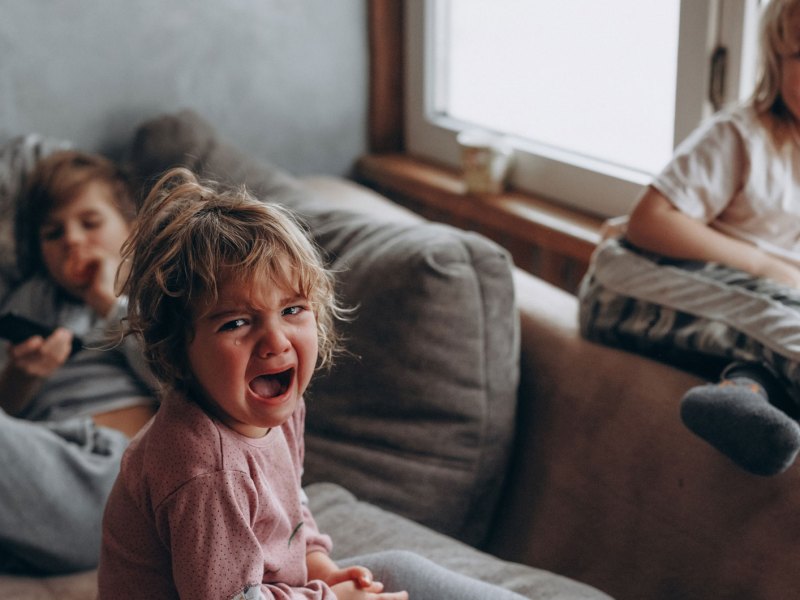 Drei Kinder auf dem Sofa. Das Gro&szlig;e im Hintergrund h&auml;lt die Fernbedienung, eines sitzt auf der Lehne und lacht und das dritte, vermeintlich j&uuml;ngste, sitzt vorn auf dem Sofa und schreit und weint.