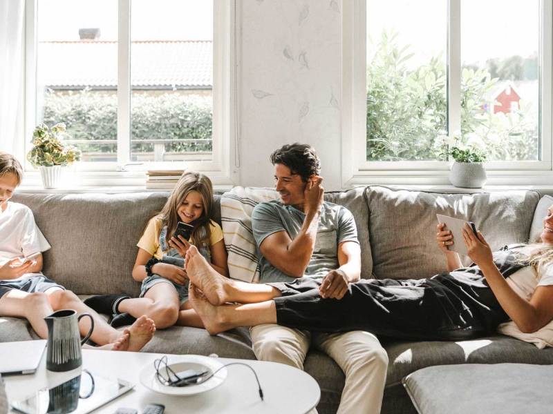 Vierk&ouml;pfige Familie sitzt auf dem Sofa, alle haben ein digitales Ger&auml;t in den H&auml;nden.