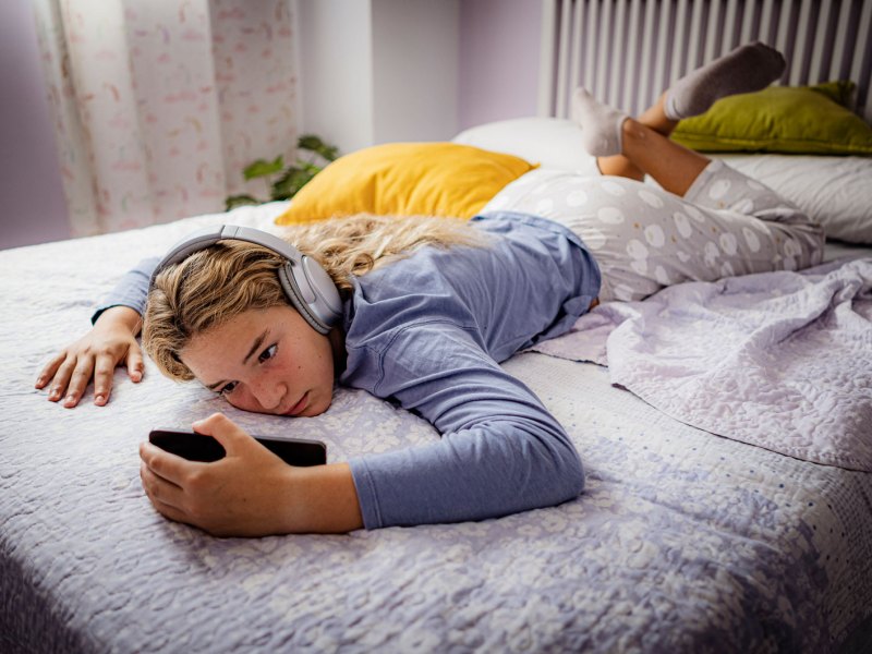 M&auml;dchen im Teenageralter, liegt mit ihrem Handy und Kopfh&ouml;rern faul auf ihrem Bett.
