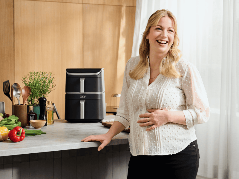 Junge blonde Frau steht am Tisch, neben ihr eine Heißluftfritteuse