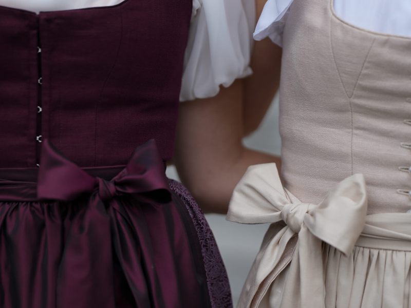 Zwei Frauen im Dirndl stehen eng nebeneinander, die Schleifen ihrer Sch&uuml;rzen sorgf&auml;ltig vorne gebunden.