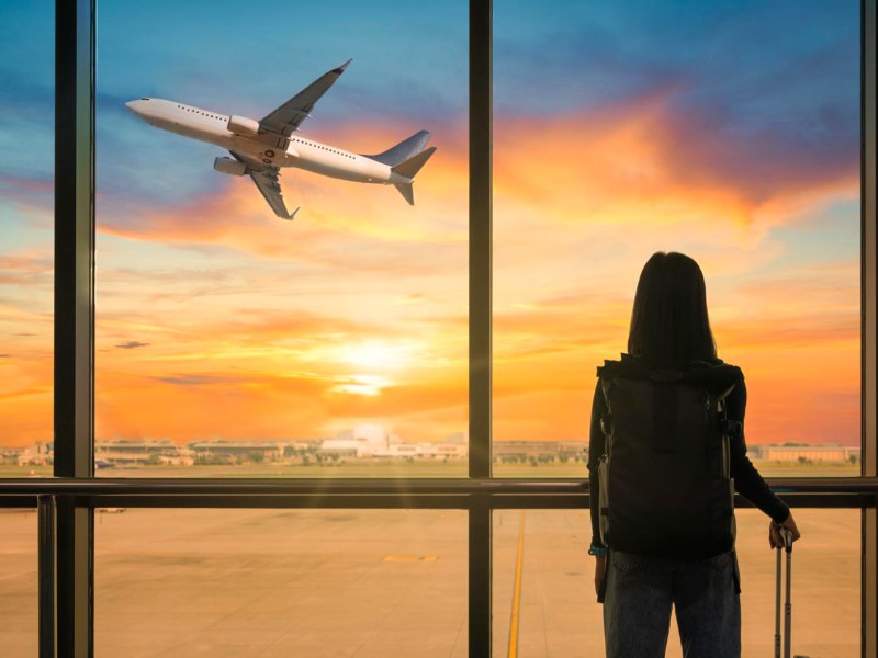 Frau steht mit Koffer am Flughafen und beobachtet aufsteigendes Flugzeug bei Sonnenuntergang
