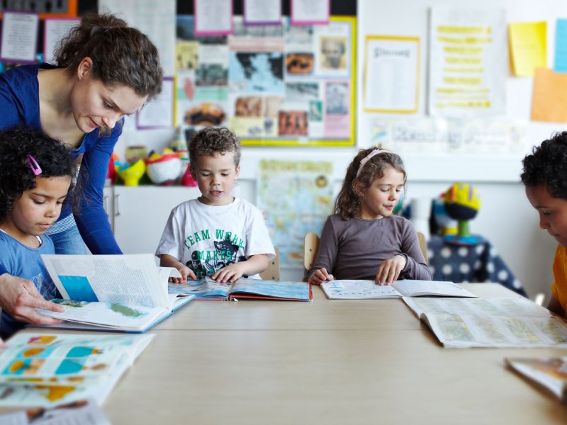 Gruppe Erstkl&auml;ssler sitzt mit B&uuml;chern an einem gro&szlig;en, runden Tisch, die Lehrerin beugt sich &uuml;ber eines der Kinder, um zu helfen.