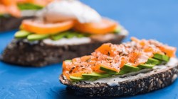 Sandwiches mit ger&auml;uchertem Lachs, Eiern, Kernen, Frischk&auml;se und Avocado auf blauem Hintergrund.