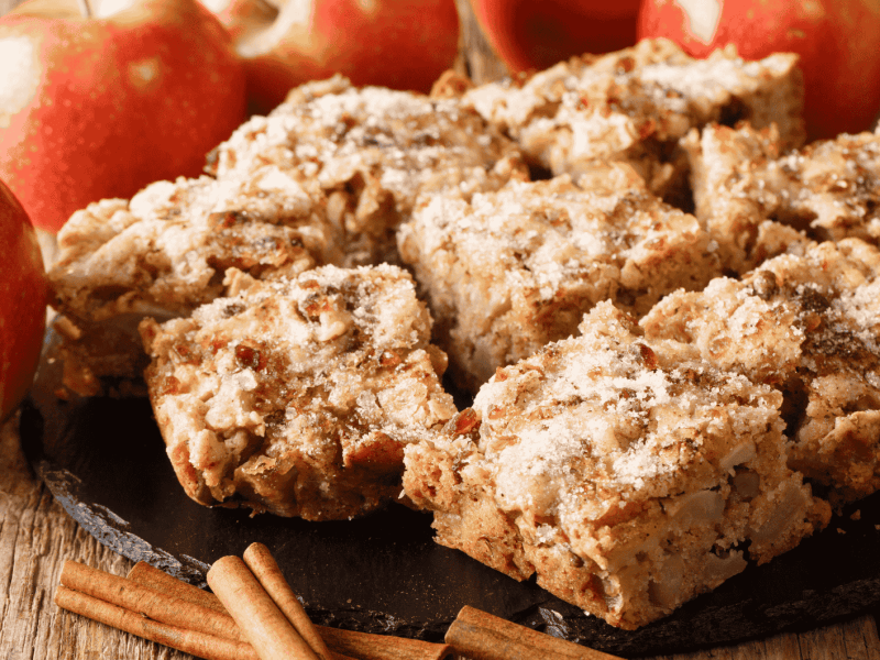 Apfel-Zimt-Kuchen auf einem Holztisch neben &Auml;pfeln und Zimtstangen.