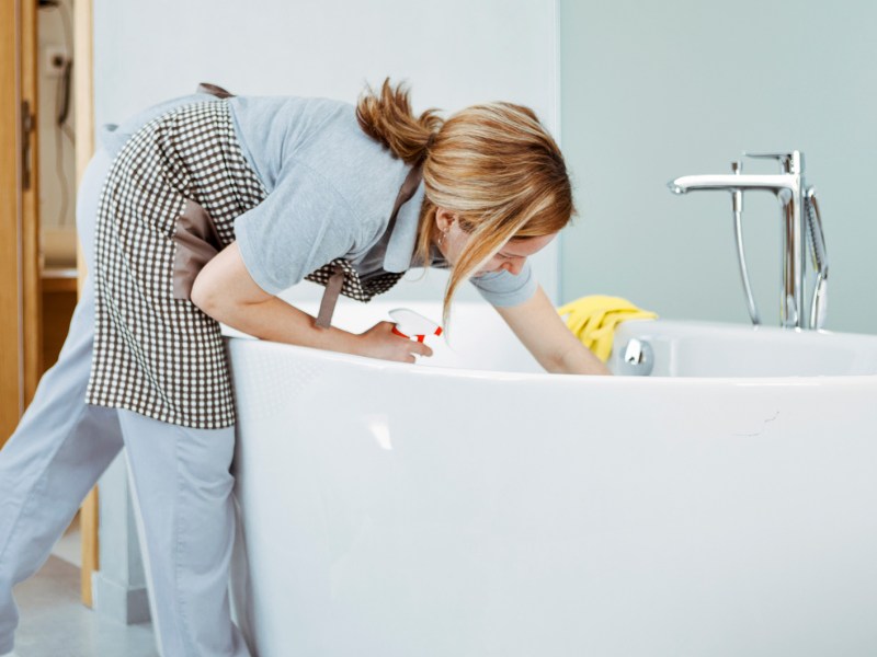 Frau putzt frei stehende Badewanne und trägt eine Schürze.