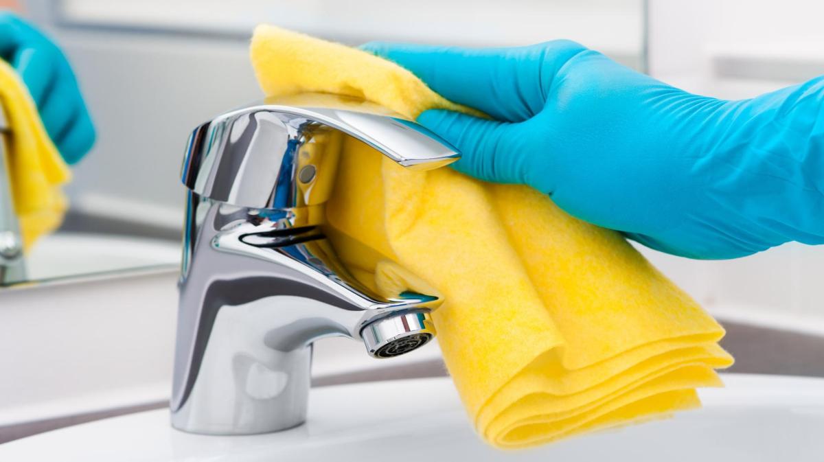 Eine Hand mit einem blauen Handschuh reinigt den Wasserhahn am Waschbecken mit einem gelben Tuch.