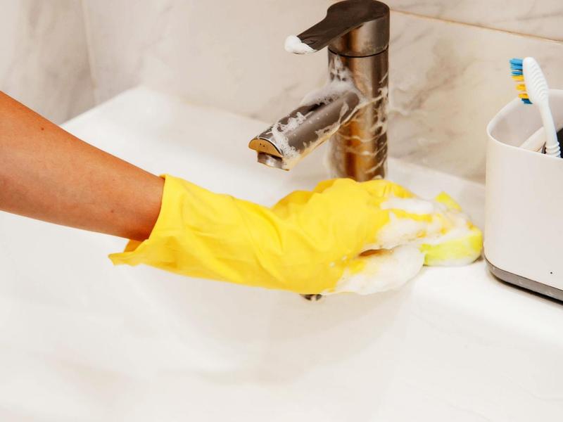 Frau mit gelben Handschuhen reinigt ein Waschbecken aus Keramik.