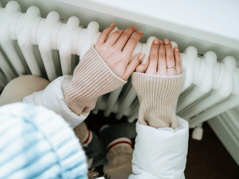 Frau sitzt warm angezogen vor der Heizung und h&auml;lt ihre H&auml;nde auf die Heizung.