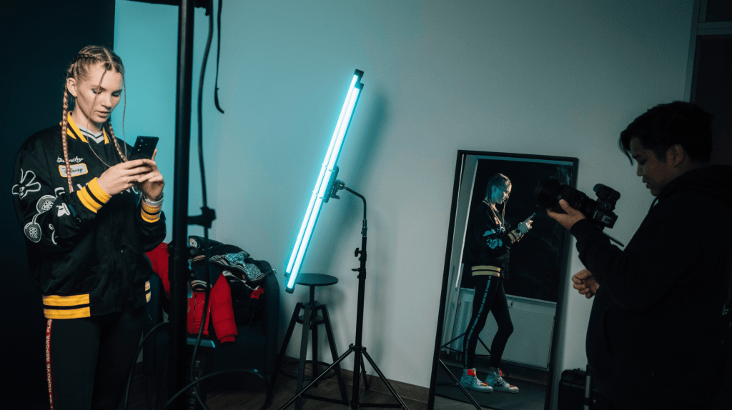 DJ LUNAX steht mit Handy in einem Raum und wird von Kameramann fotografiert.