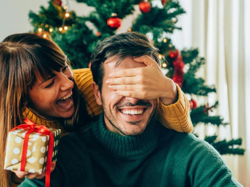 Frau hält Mann vor dem Weihnachtsbaum die Hand vor die Augen, um ihn mit einem Geschenk zu überraschen.