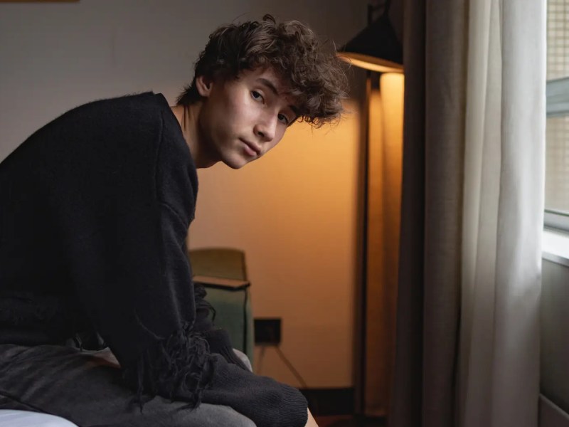 Porträt eines Teenagers: 16-jähriger Junge sitzt auf einem Bett in einem dunklen, loftartigen Zimmer gegenüber eines Fensters.