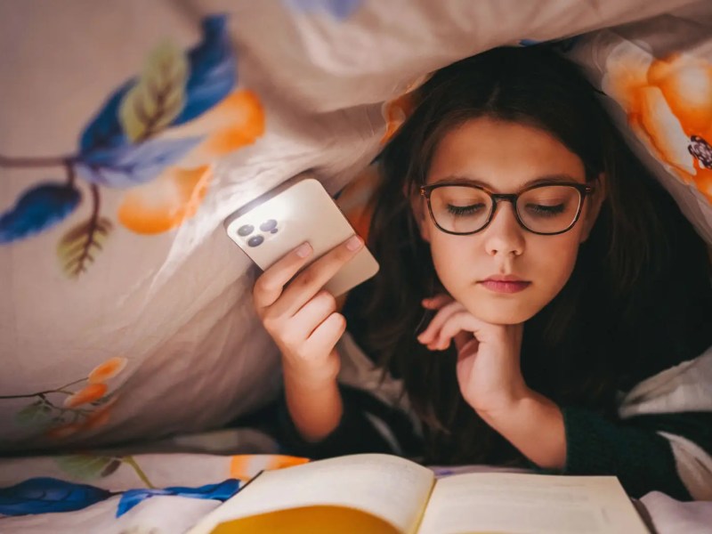 Teenager-Mädchen unter der Bettdecke, macht sich Licht mit dem handy und liest ein Buch.