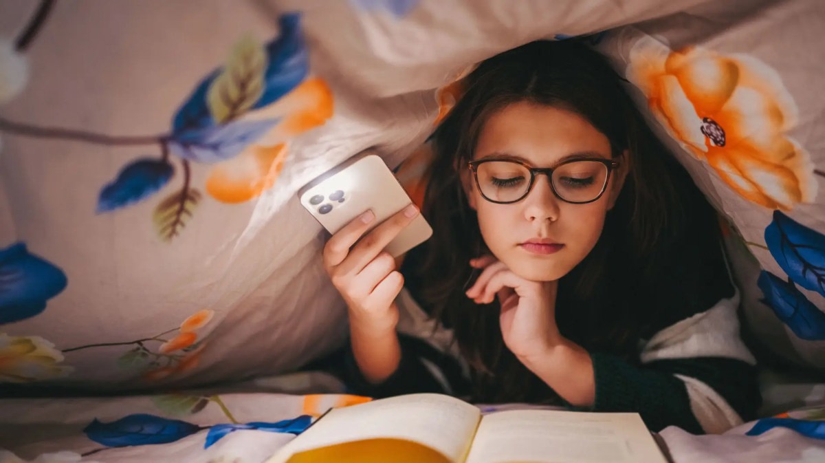 Teenager-Mädchen unter der Bettdecke, macht sich Licht mit dem handy und liest ein Buch.