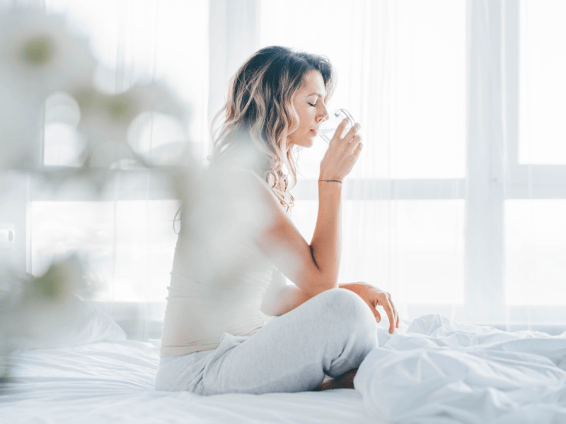 Eine gelassene junge Frau sitzt auf einem weißen Bett und trinkt vor einem hellen Fenster ein Glas Wasser. Das Konzept von morgendlichem Wohlbefinden, gesunden Gewohnheiten und einem frischen Start in den Tag.