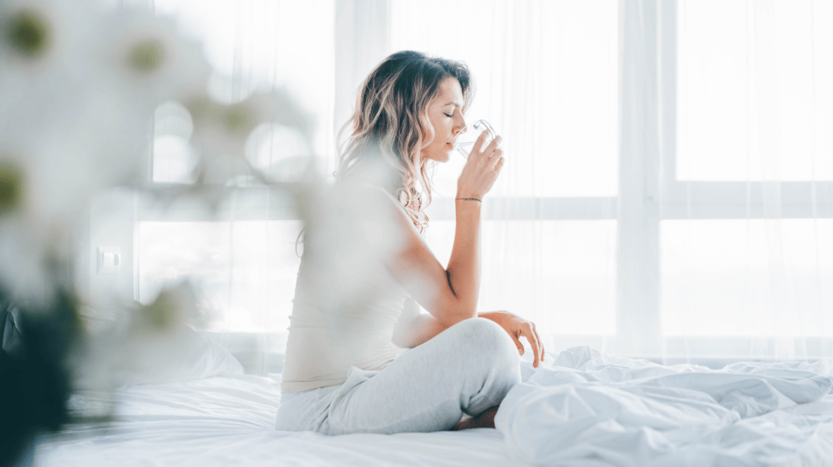 Eine gelassene junge Frau sitzt auf einem weißen Bett und trinkt vor einem hellen Fenster ein Glas Wasser. Das Konzept von morgendlichem Wohlbefinden, gesunden Gewohnheiten und einem frischen Start in den Tag.