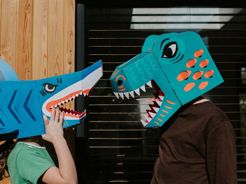 Geswister tragen Haifisch- und Dino-Maske und stehen sich gegen&uuml;ber.