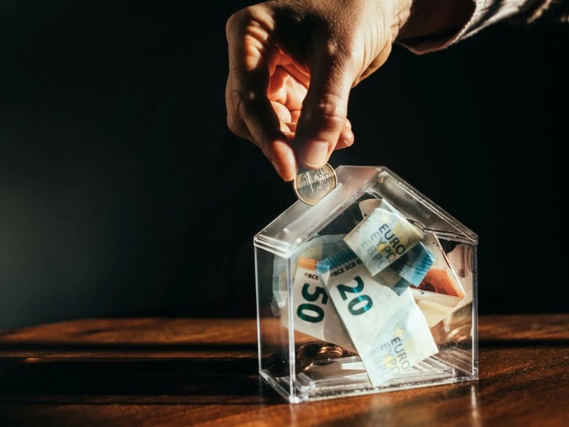 Frau wirft Geld in eine transparente Spardose, die wie ein Haus aussieht.